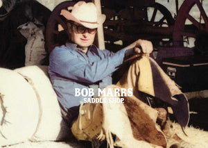 Bob Marrs American Cowboy and Saddle Maker wearing chaps along side his bedroll and chuckwagon on the range